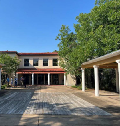 A sunny view of a modern building with a red-tiled roof, surrounded by greenery and outdoor gathering space.
