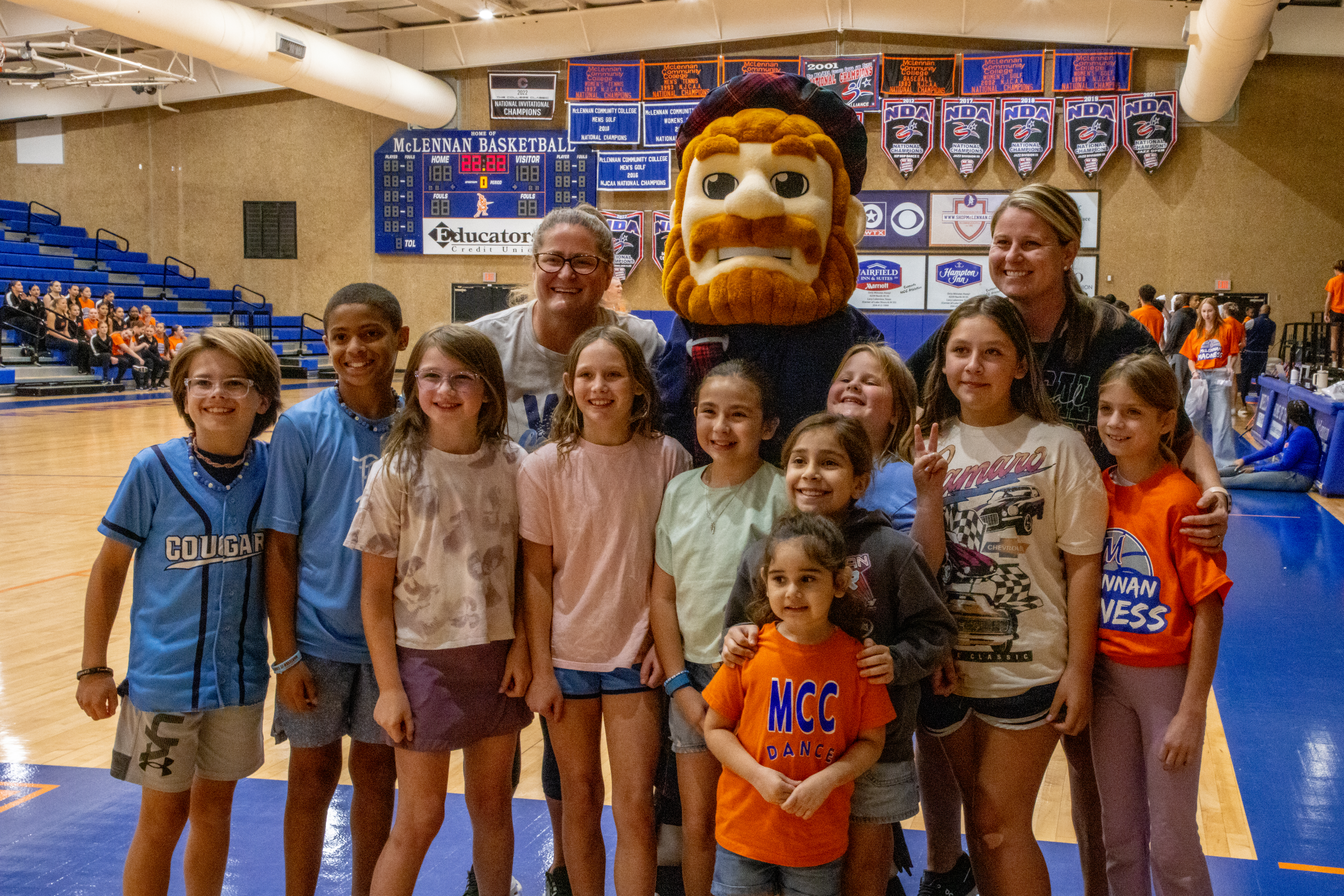 A group of smiling children and adults pose with the McLennan Community College Highlander mascot in a gymnasium. 
