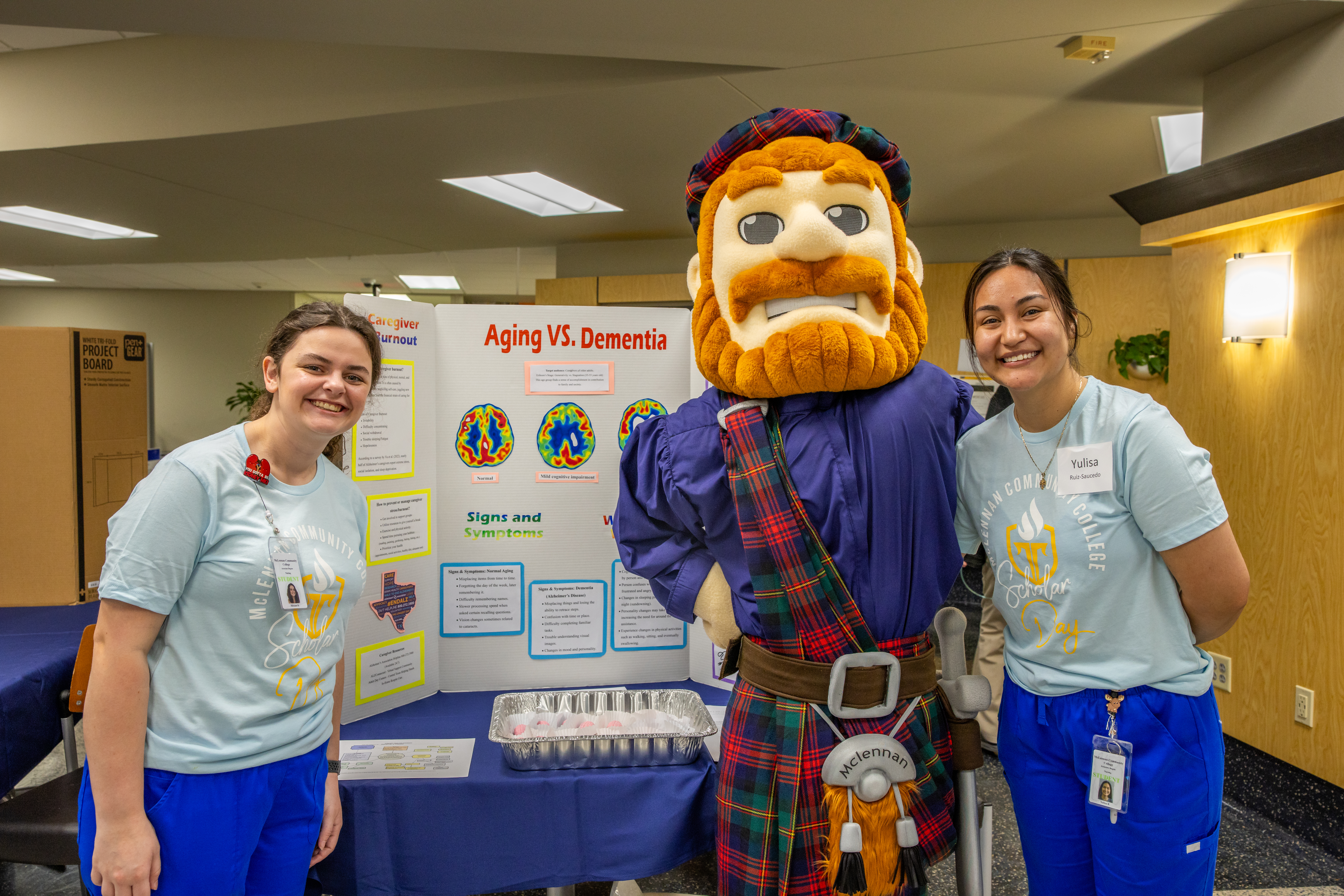 Two McLennan Community College nursing students stand smiling with the Highlander mascot in front of a presentation board titled “Aging vs. Dementia.”