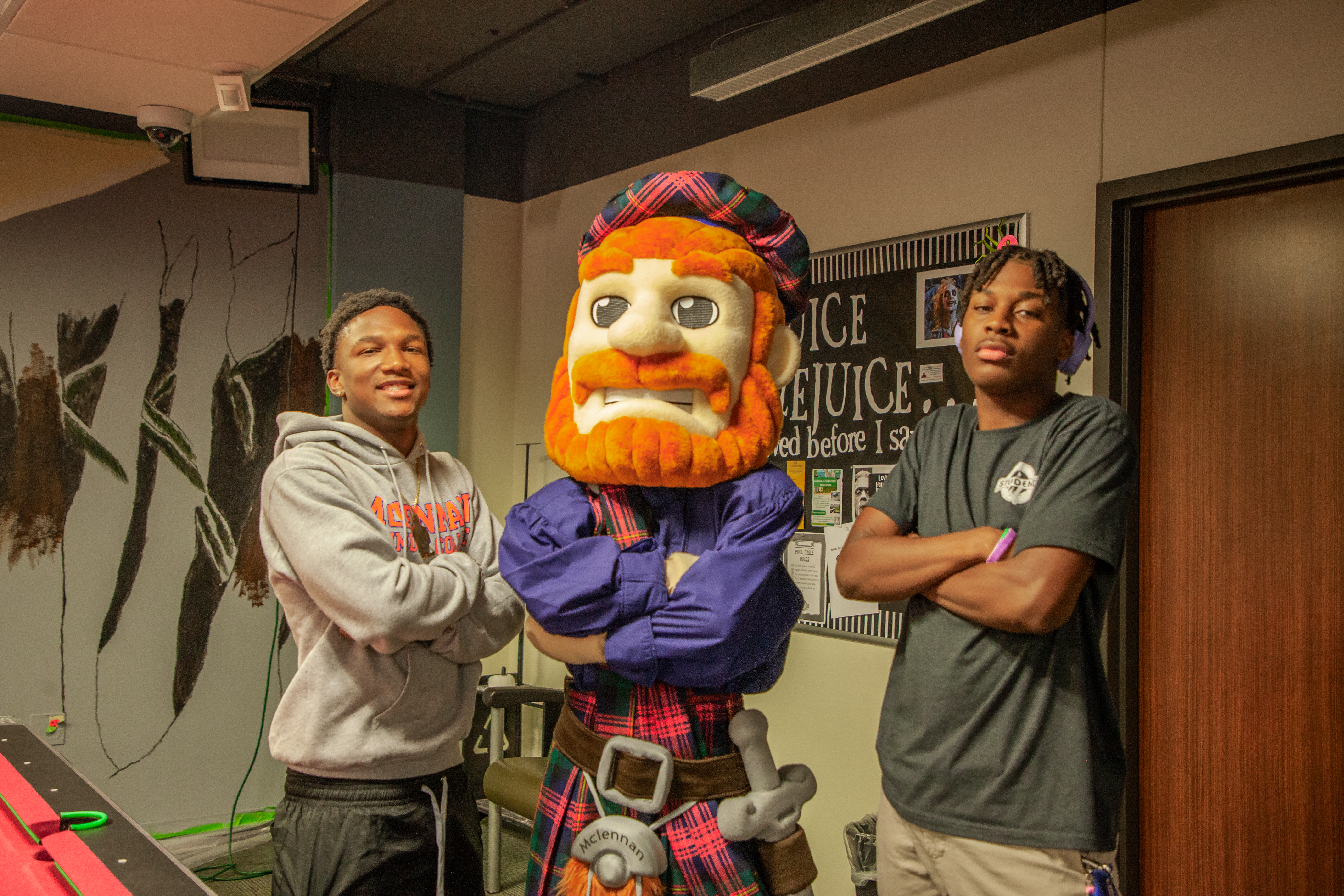 Two McLennan Community College students pose with the Highlander mascot, all with arms crossed, in a student lounge with bulletin boards and wall art.