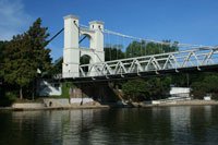 A suspension bridge spans over a calm river, surrounded by lush greenery and blue skies.