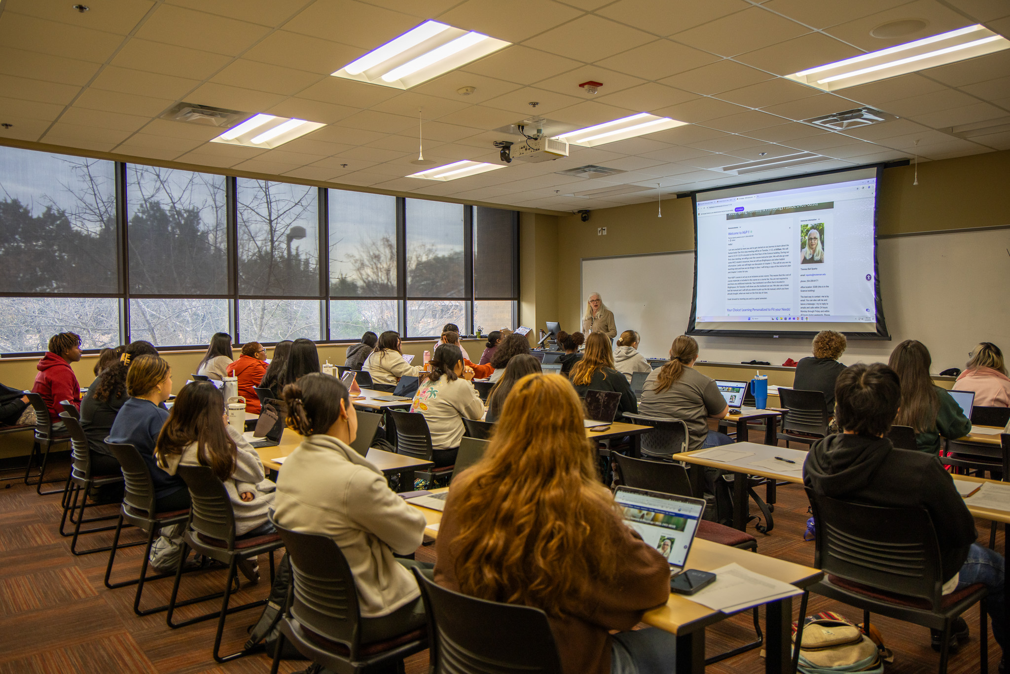 A wide-angle, indoor shot of a brightly lit university classroom filled with students seated at long wooden desks, facing a large projector screen at the front. The students, mostly seen from the back or side, are using laptops and notebooks. An instructor with long white hair stands near the front, next to the screen which displays a "Welcome to AUPP" orientation page featuring her photo and contact information.  Large windows on the left wall reveal bare trees under an overcast sky. The room features a drop ceiling with recessed fluorescent lighting and a dark-patterned carpet. The overall atmosphere is one of focused academic engagement.