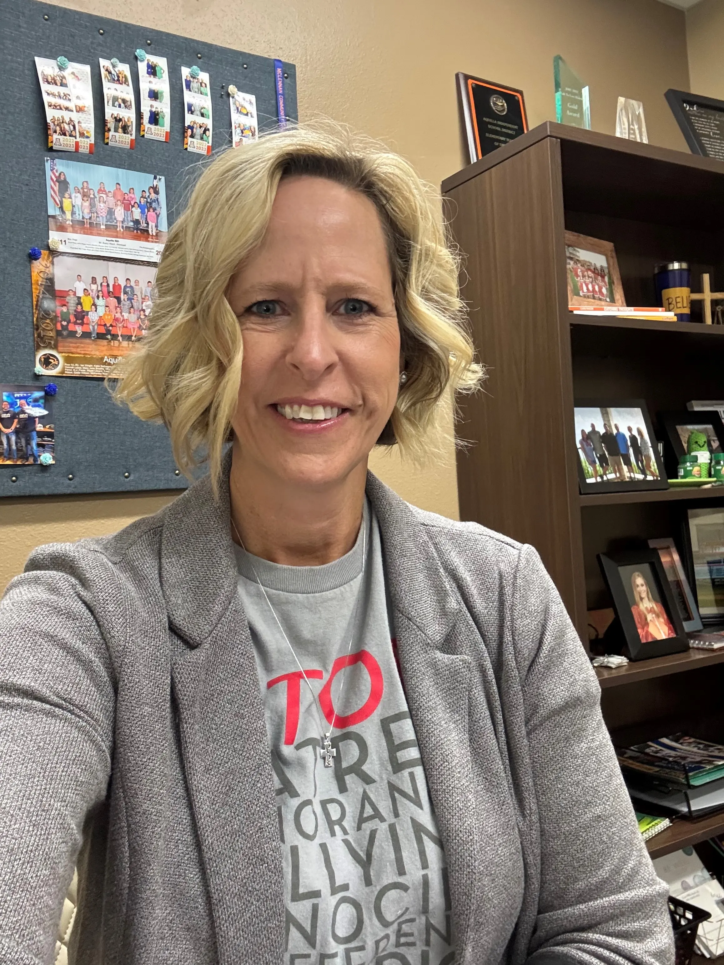 Smiling woman taking a selfie in an office, with a bulletin board of photos and a bookshelf with awards and framed pictures in the background.