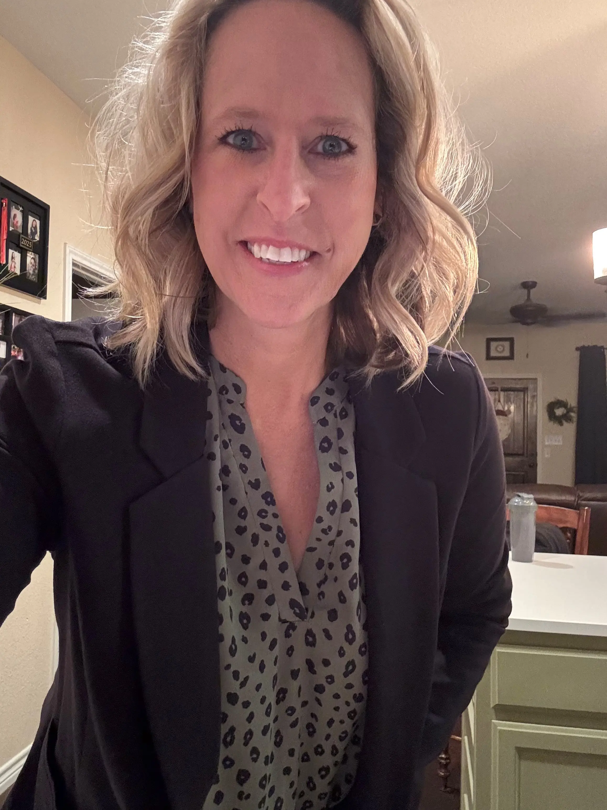 Professional headshot of a woman with short, curly blonde hair, smiling in an office setting with a bulletin board and framed photos in the background.