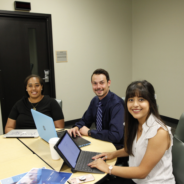 Three people sit around a conference table in an office setting, working on laptops and reviewing printed materials. Brochures, notebooks, and a coffee cup are on the table. A closed door and wall signage are visible in the background.