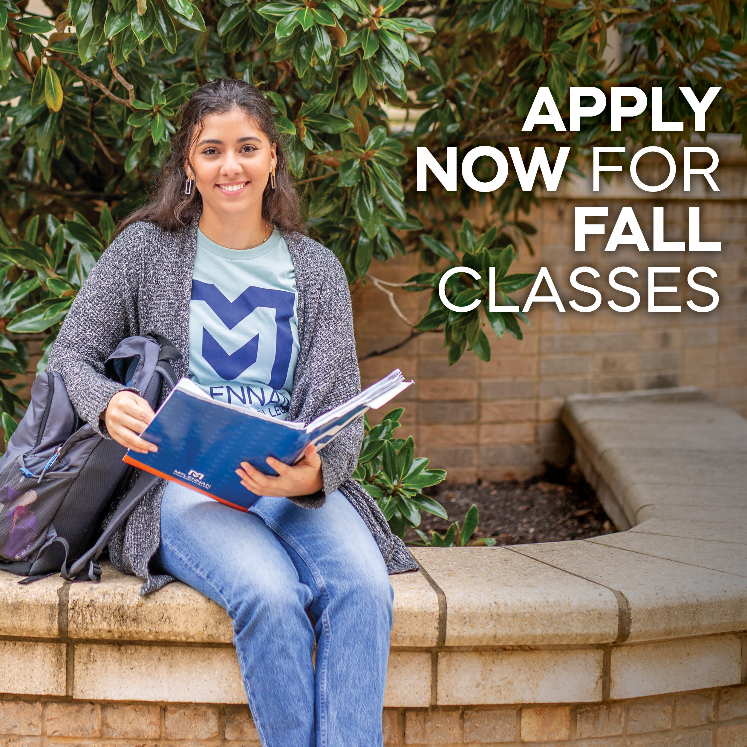 A student wearing a light blue MCC T-shirt and gray cardigan sits on a stone bench outdoors, writing in an open book with a pen. A black backpack rests nearby. Green foliage and a brick wall are in the background. Text on the image reads: “APPLY NOW FOR FALL CLASSES.”