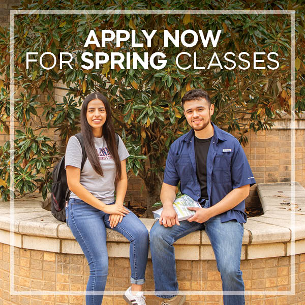 Two students sitting on a curved stone bench outdoors in front of a brick wall and leafy green plants. One student is wearing a gray T-shirt and jeans with a backpack, while the other is wearing a dark blue button-up shirt over a black T-shirt and jeans, holding a campus map. Text above them reads: “APPLY NOW FOR SPRING CLASSES.”