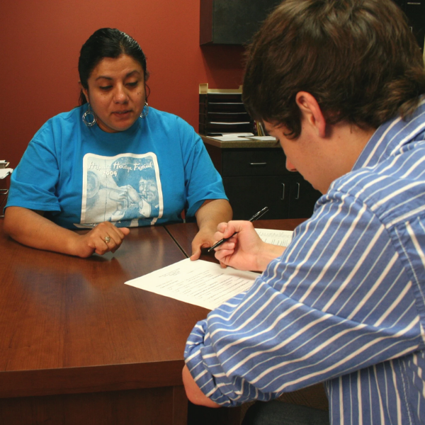 Two people sit across from each other at a wooden desk in an office setting. One person, wearing a blue shirt with a printed graphic, rests their hands on the table. The other person, wearing a striped shirt, is writing on a form or document. The background includes office furniture and shelves.