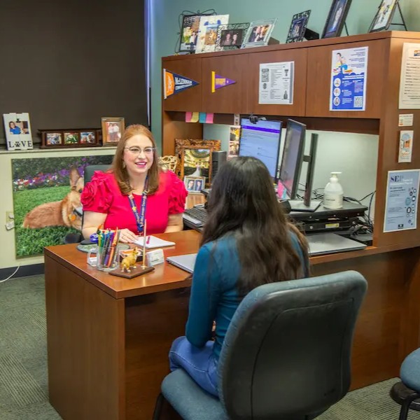 A student sits across from a school counselor in an office. The counselor sits behind a wooden desk with two monitors, office supplies, and hand sanitizer. The student holds a notebook while listening. The office walls and shelves display personal photos, posters, and college pennants, creating a supportive academic setting.