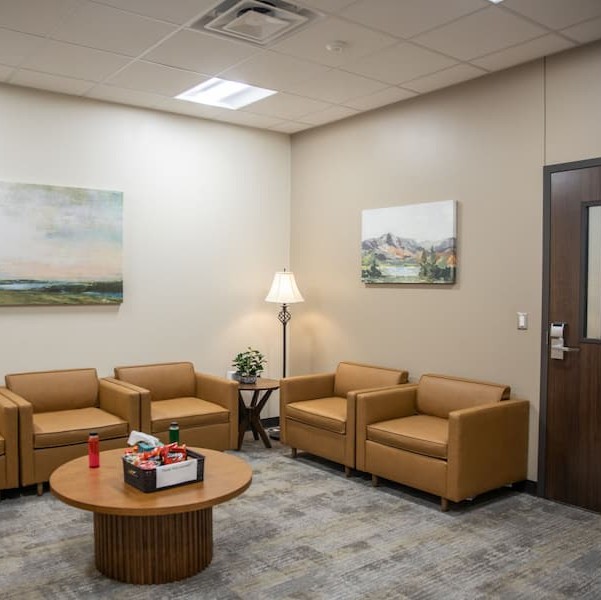 Welcoming waiting area with comfortable tan armchairs, a round coffee table, floor lamp, and landscape artwork on the walls