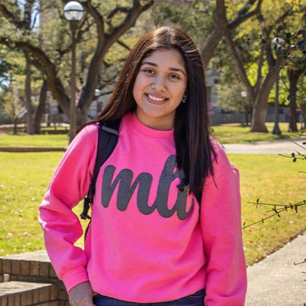 Person standing in a park-like setting wearing a bright pink sweatshirt with 'mlb' in large dark gray letters, carrying a black backpack. Background includes green grass, trees, a pathway, benches, and light posts.
