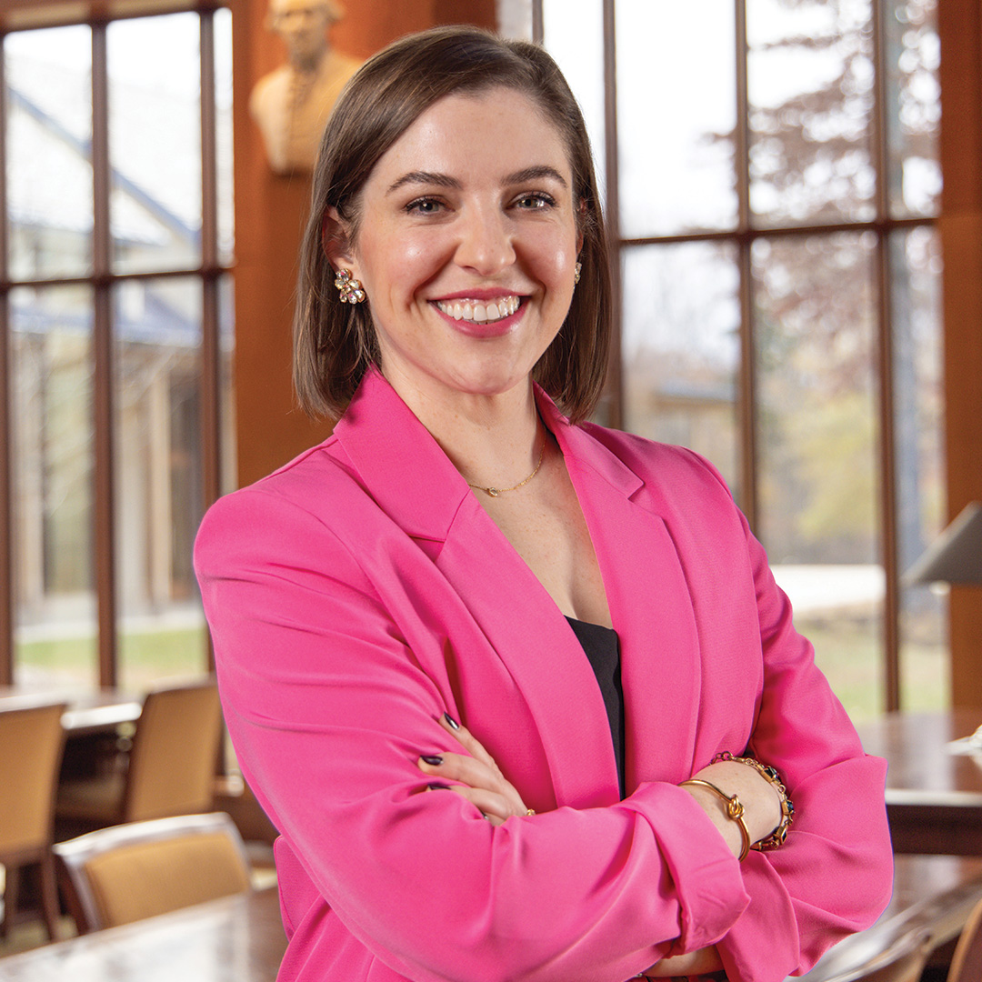 Smiling woman in a bright pink blazer with arms crossed, standing indoors in front of large windows.