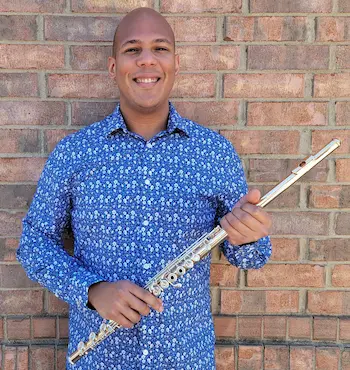 Smiling person in a blue patterned shirt holding a silver flute in front of a brick wall.