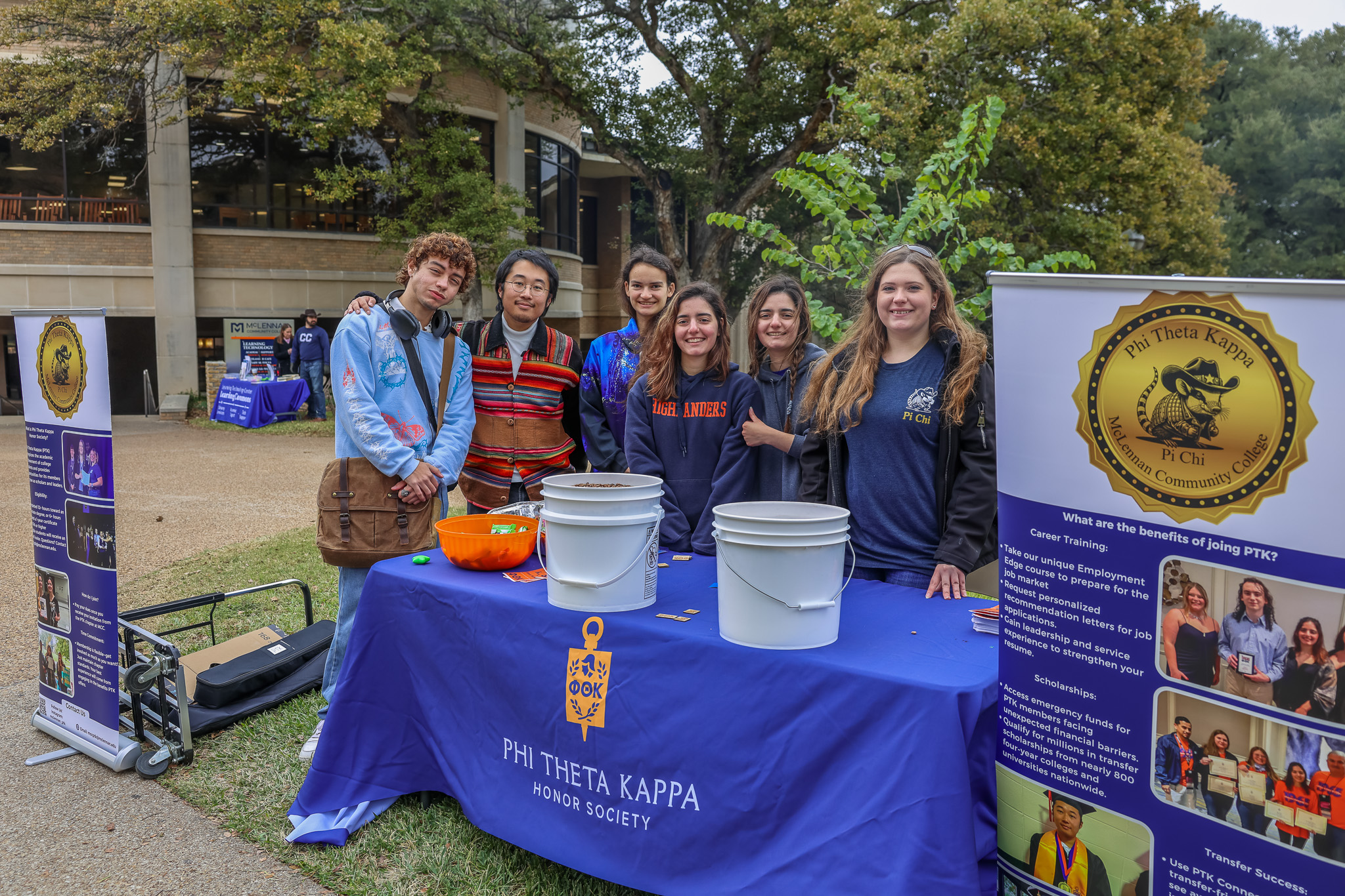 Phi Theta Kappa students huddled together to pose for a picture behind a Phi Theta Kappa table cloth, displays, buckets of uncooked beans, and candy.