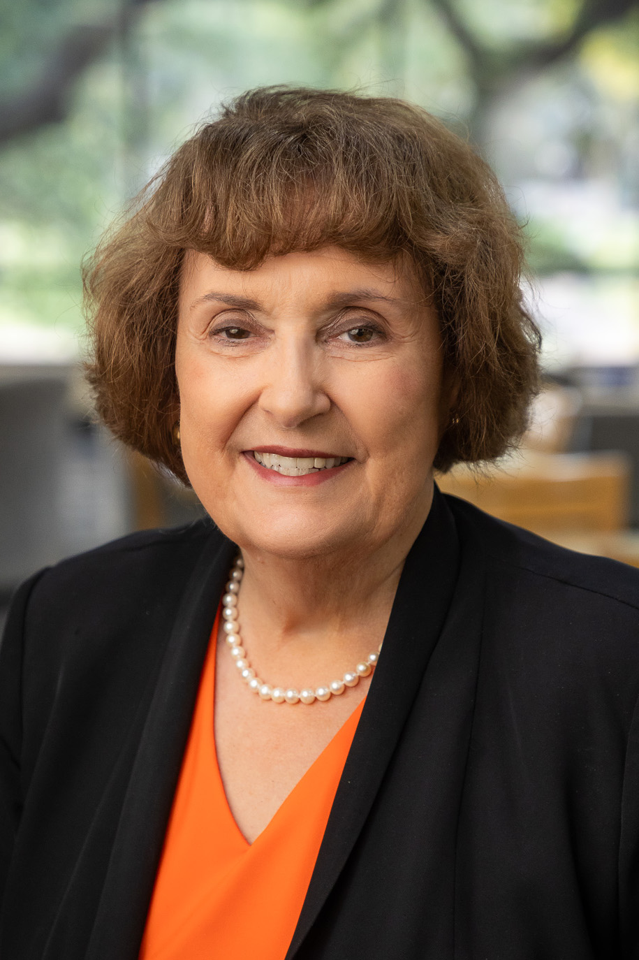 Smiling woman in a black blazer and orange top, wearing a pearl necklace, in an indoor setting with a softly blurred background.