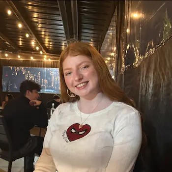 Smiling young woman with long red hair wearing a white shirt with a Spider-Man logo, seated indoors under string lights.