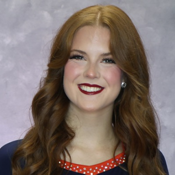 Smiling red-haired woman with wavy hair, red lipstick, and pearl earrings against a gray studio backdrop.