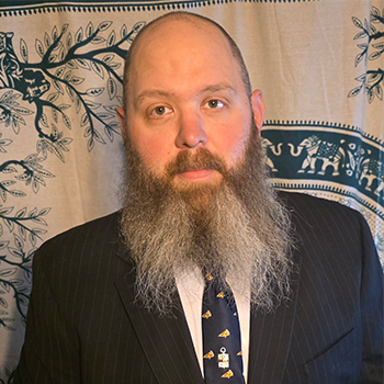 A man with a full beard and mustache wearing a suit and tie, standing in front of a patterned backdrop.