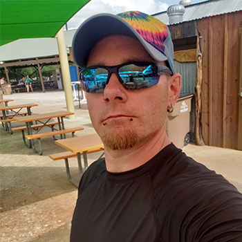 Person wearing a colorful cap and reflective sunglasses taking a selfie outdoors near picnic tables.