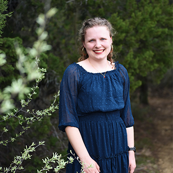 A smiling young woman in a blue dress stands outdoors surrounded by greenery.