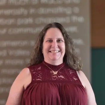 Smiling woman with long wavy hair wearing a burgundy sleeveless top, standing indoors in front of a blurred presentation screen.