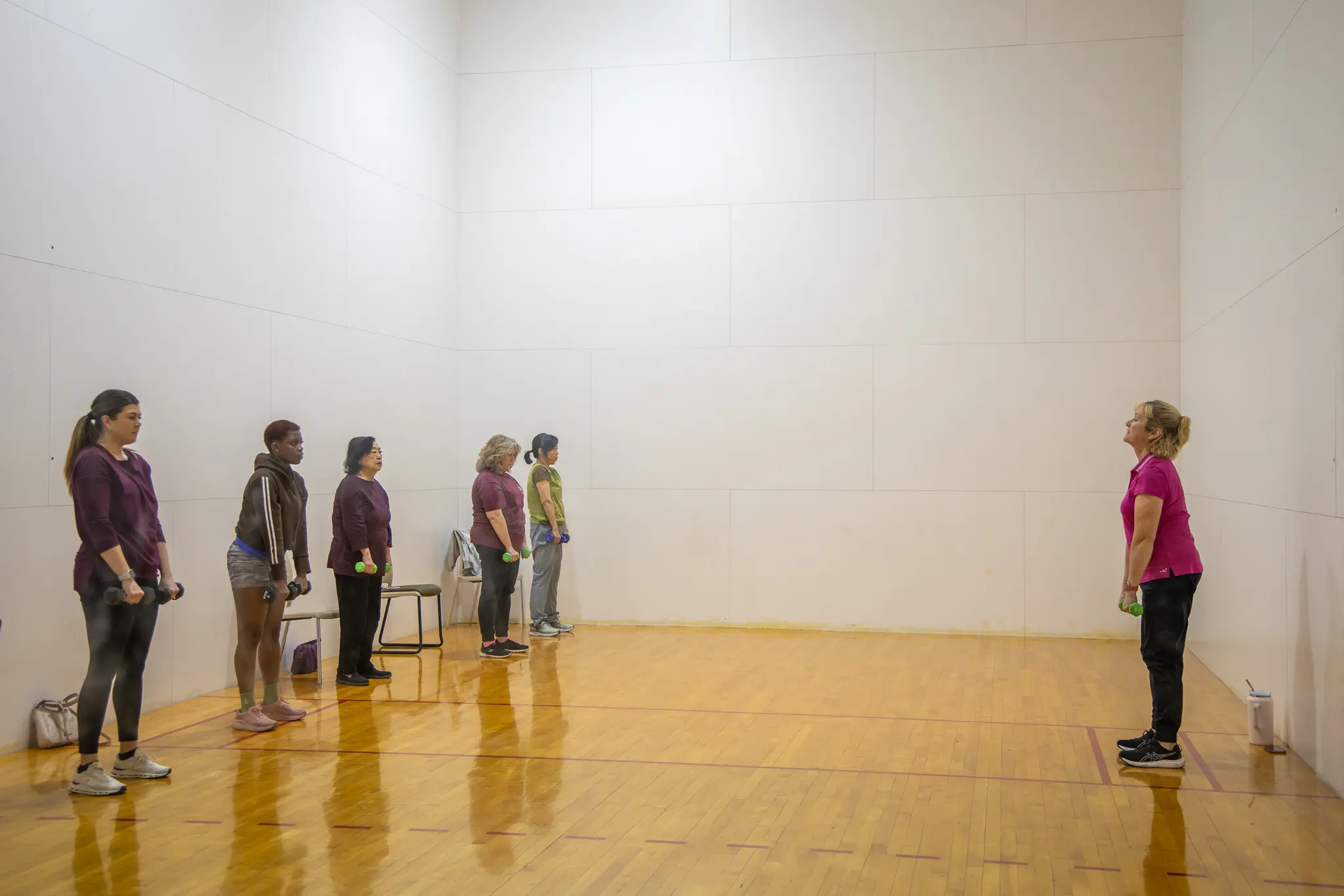Group of women in a fitness class standing in a line, each holding weights, in a gymnasium setting.
