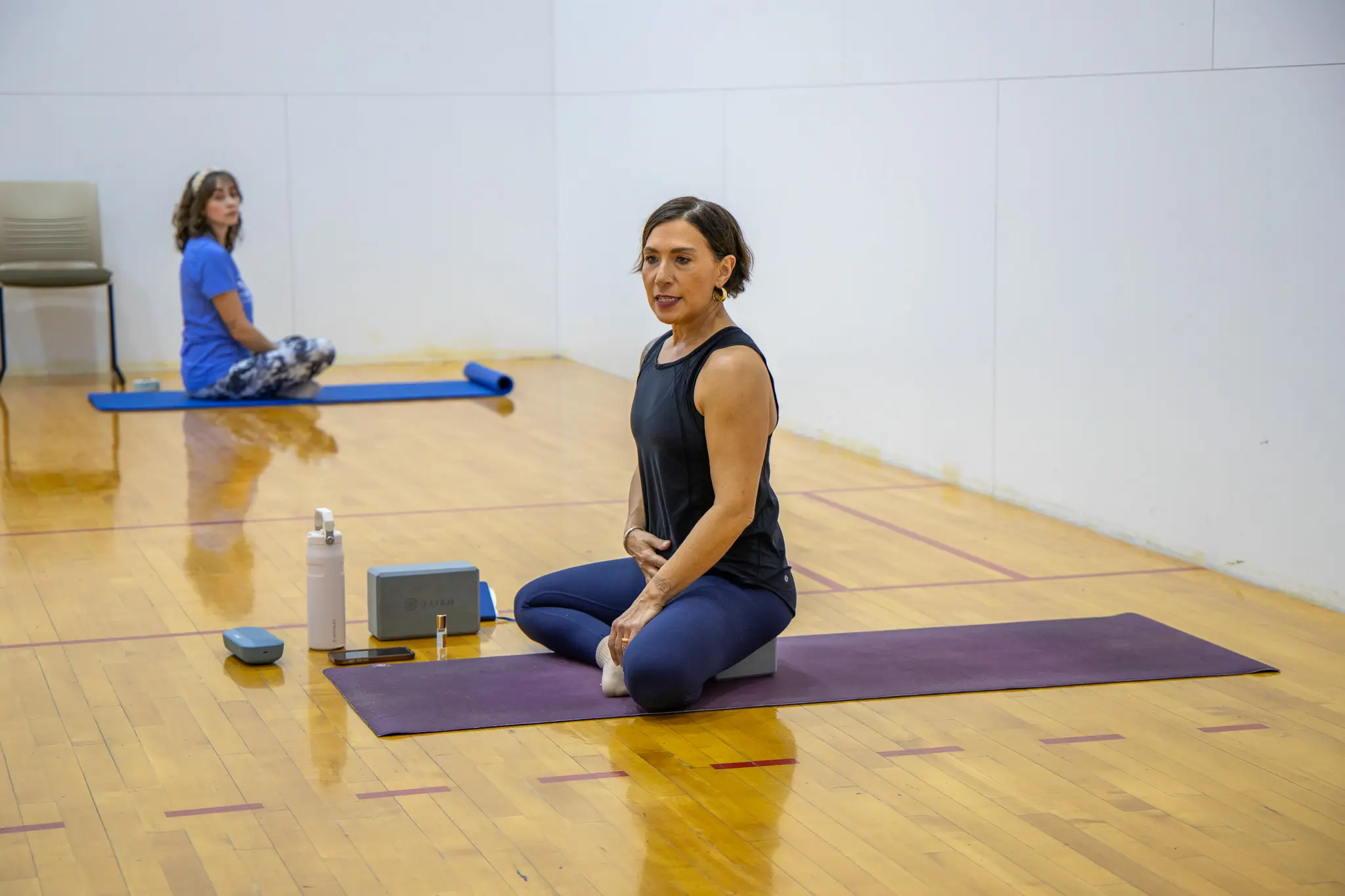 A yoga instructor demonstrates a seated pose in a gym setting while a student practices in the background.